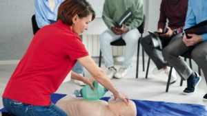 Una mujer en primer plano practicando técnicas de reanimación cardiopulmonar básica usando desfibrilador en caso de emergencia sobre un muñeco de práctica y un grupo de estudiantes sentados al fondo